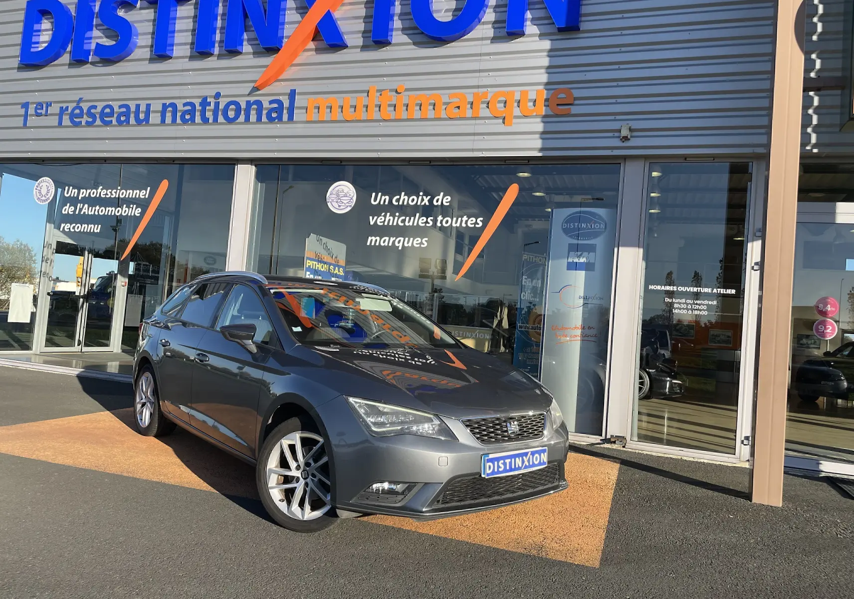 SEAT LEON ST gris en 3/4 avant droit, stationnée devant un showroom avec plaques Distinxion visibles.