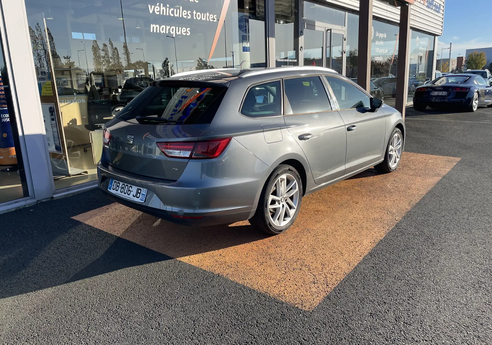 SEAT LEON ST gris en 3/4 arrière droit, garée devant une vitrine avec reflets et plaques françaises visibles.