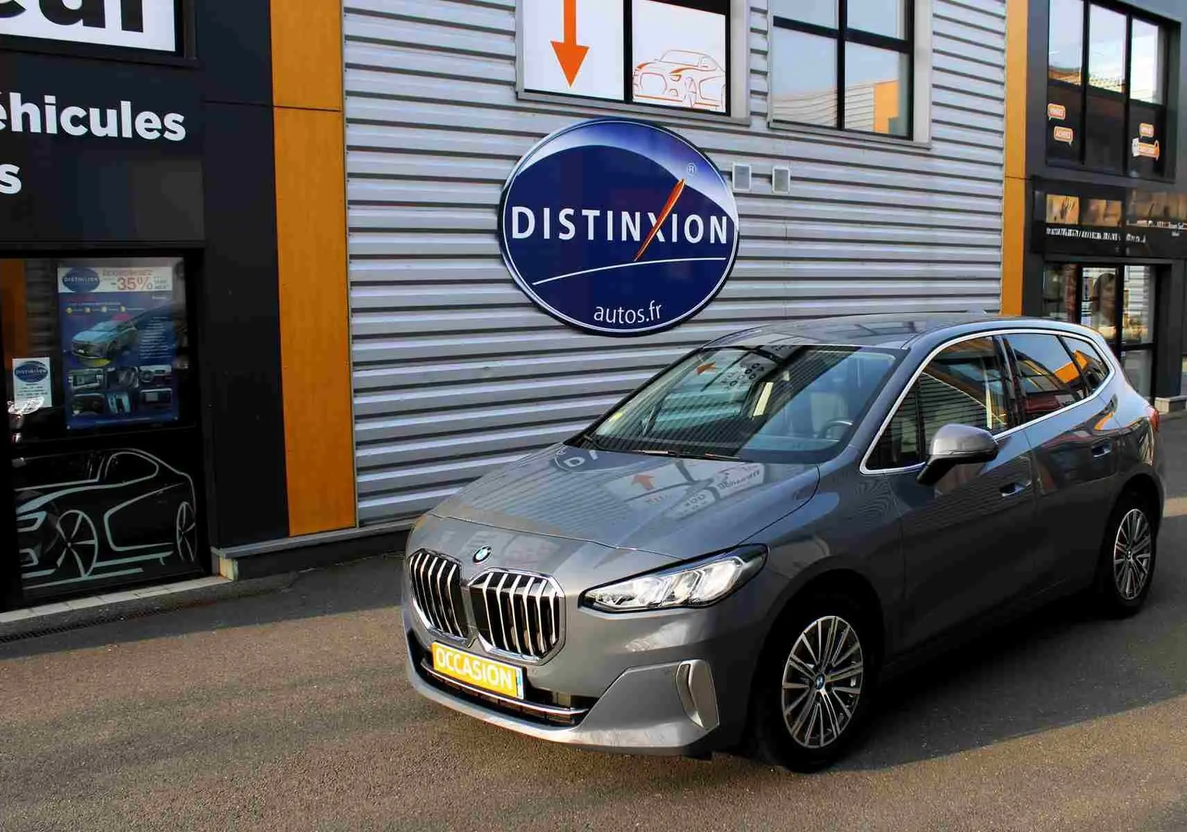 BMW Série 2 Active Tourer gris Nardo, vue 3/4 avant droit, garée devant un bâtiment commercial.