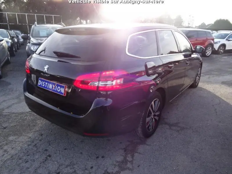 Peugeot 308 SW noire vue en 3/4 arrière droit, avec feux arrière allumés sur parking extérieur ensoleillé.