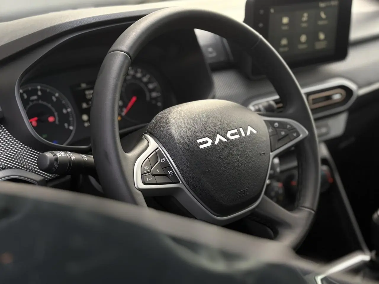 Vue rapprochée du volant noir Dacia avec commandes intégrées et tableau de bord numérique en arrière-plan.