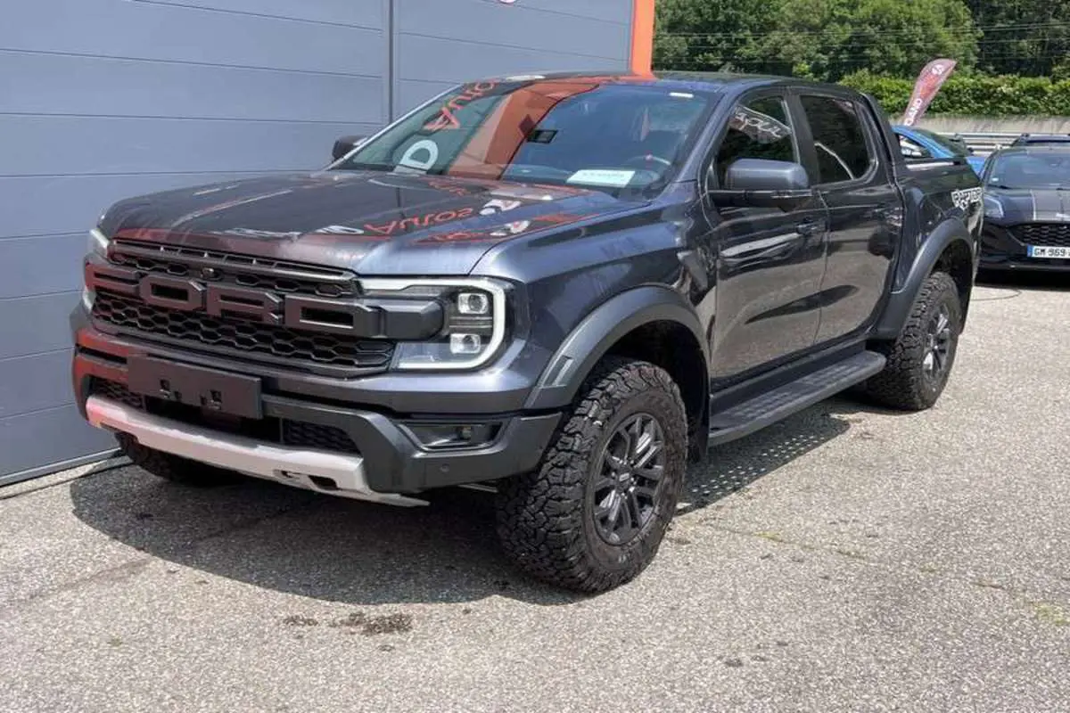 Ford Ranger Raptor 2025 gris météore en 3/4 avant droit, avec calandre imposante et pneus tout-terrain marqués.