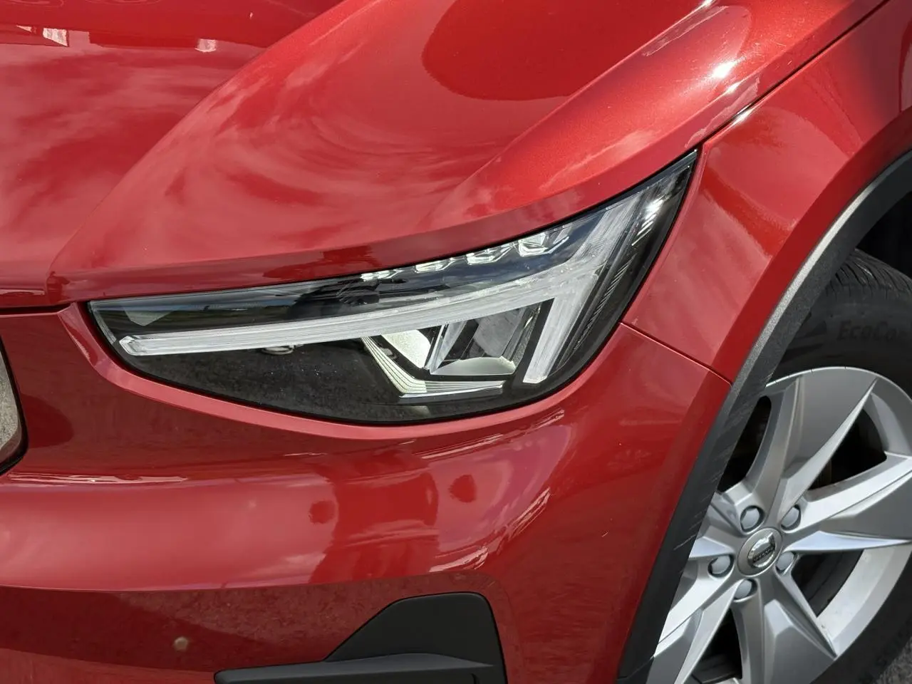 Gros plan sur le phare avant gauche et la jante alliage 18'' du Volvo XC40 rouge Fusion 2024.