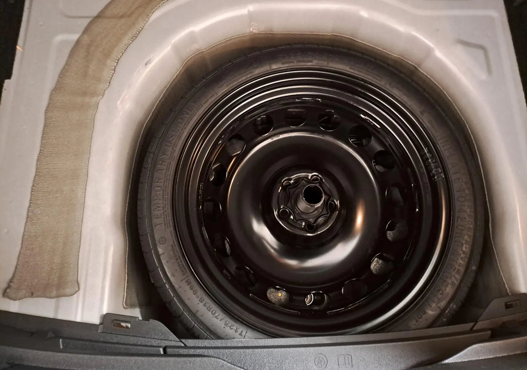 Roue de secours noire en acier dans le coffre d’un Volkswagen Taigo 2024 blanc pur, vue plongeante.