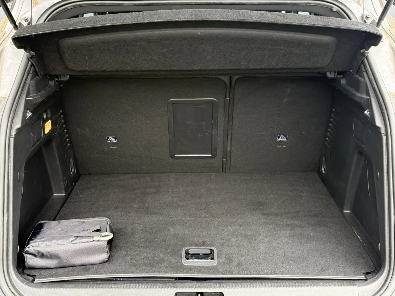 Coffre ouvert vu de l'arrière, intérieur noir avec tapis et sac de rangement, Peugeot 3008 gris Artense visible partiellement.