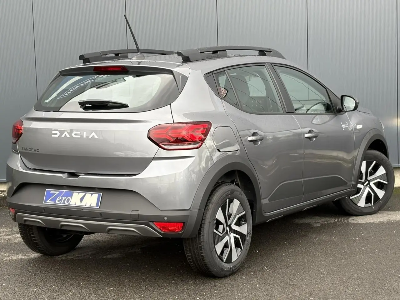 Vue 3/4 arrière droite d'une Dacia Sandero Stepway 2025 gris schiste avec barres de toit et jantes spécifiques.