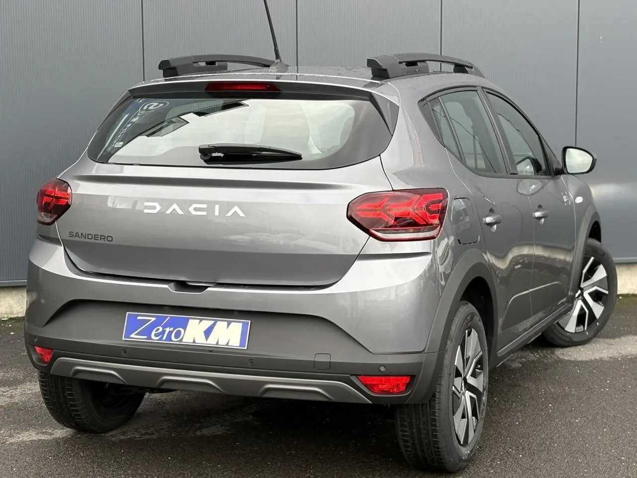 Vue 3/4 arrière droite d'une Dacia Sandero Stepway 2025 gris schiste avec barres de toit et feux arrière LED en Y.