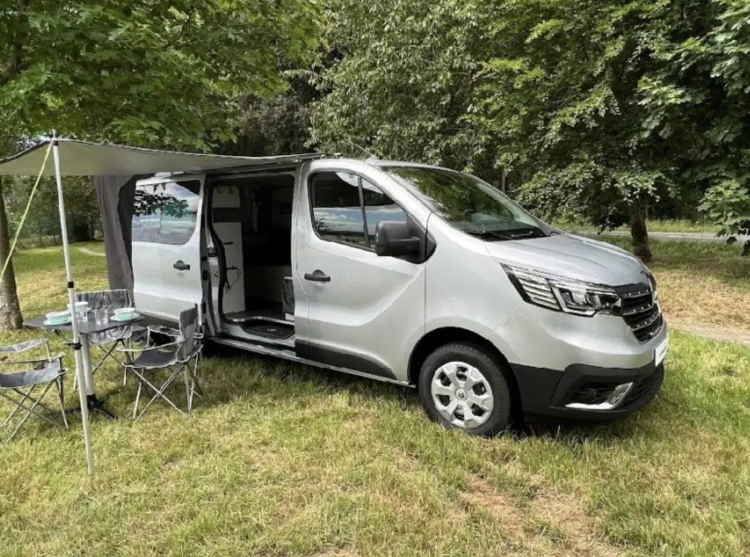 Renault Trafic Fourgon gris clair en 3/4 avant droit, porte latérale ouverte avec auvent et mobilier de camping installé.