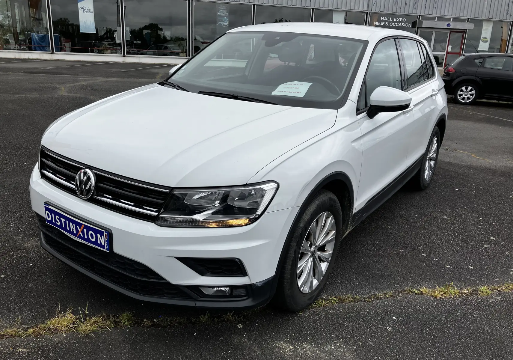 Volkswagen Tiguan blanc vu en 3/4 avant droit, avec calandre noire et feux allumés sur parking devant un hall d'exposition.