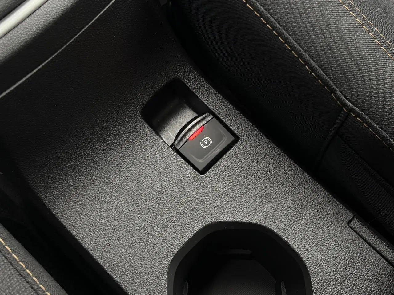 Gros plan sur le frein de parking électrique noir avec témoin rouge, entouré de sellerie gris foncé à surpiqûres contrastées.