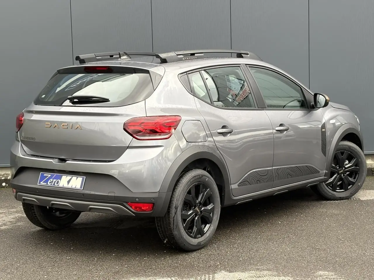 Vue 3/4 arrière droite d'une Dacia Sandero Stepway 2025 gris schiste avec jantes noires et barres de toit longitudinales.