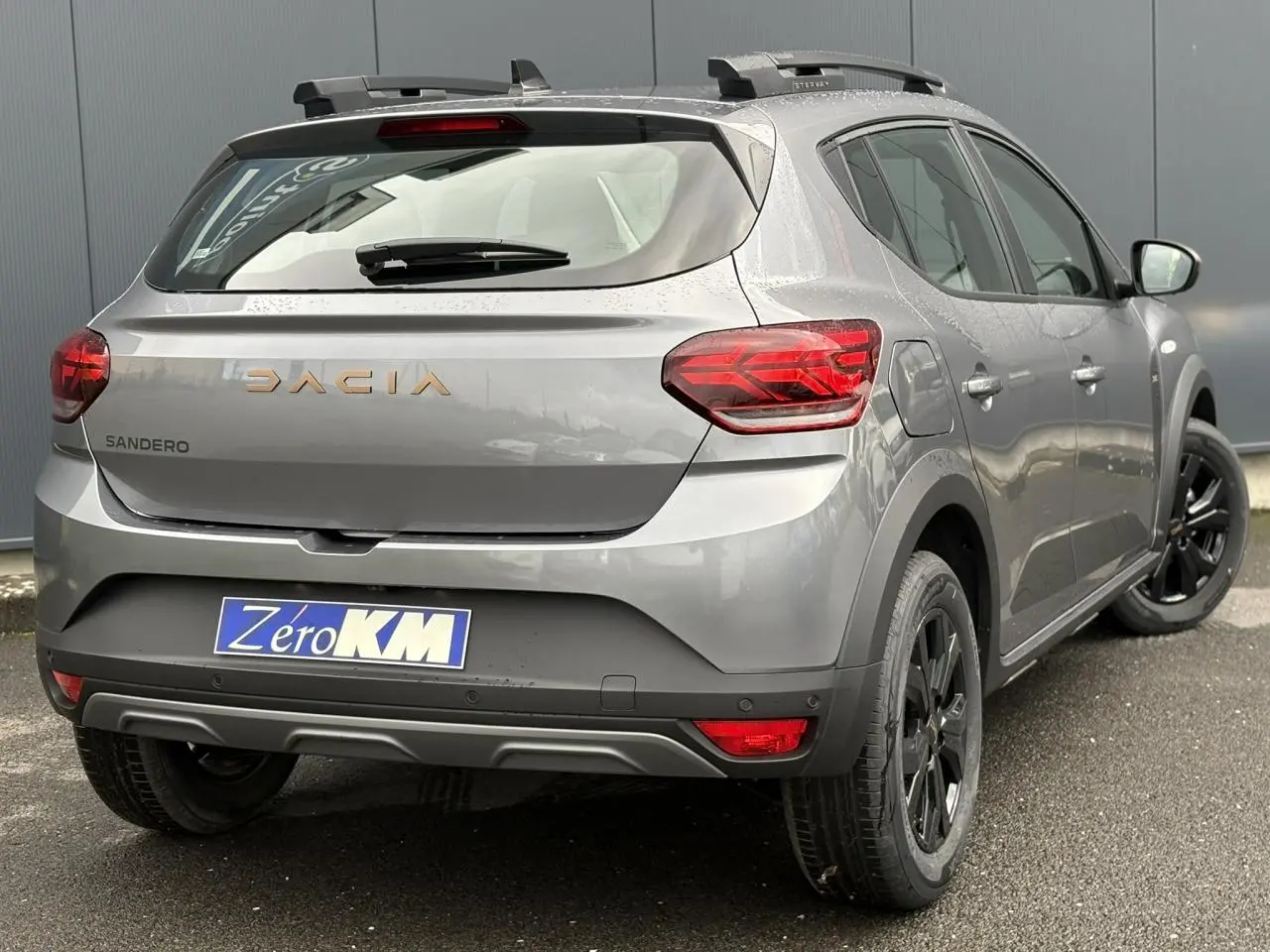 Vue 3/4 arrière droite d'une Dacia Sandero Stepway TCE 110 gris schiste avec barres de toit et jantes noires.