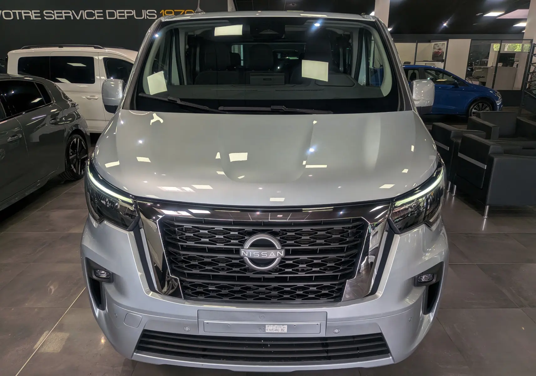 Vue frontale d'un Nissan Primastar Combi gris Highland 2026 avec calandre noire et phares LED allumés.