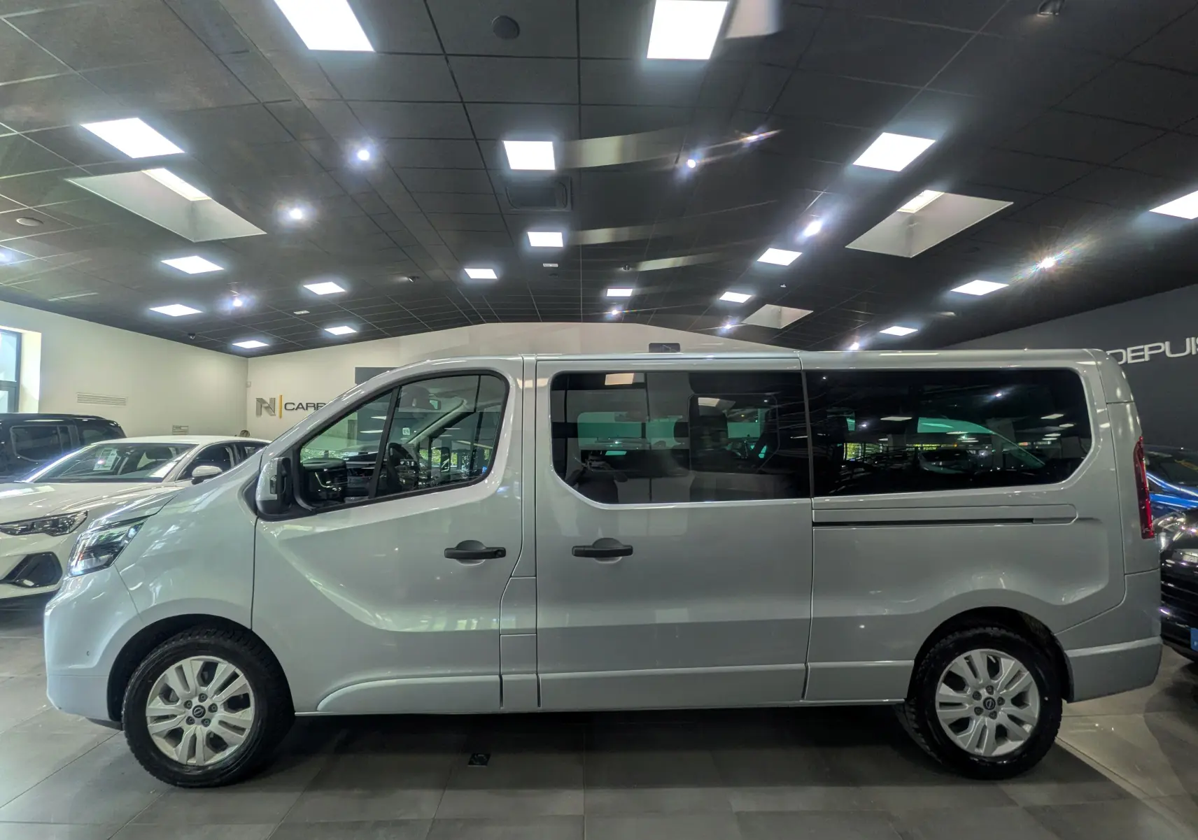 Vue de profil côté gauche du Nissan Primastar Combi gris Highland 2026 en showroom, avec vitres teintées et jantes alliage.