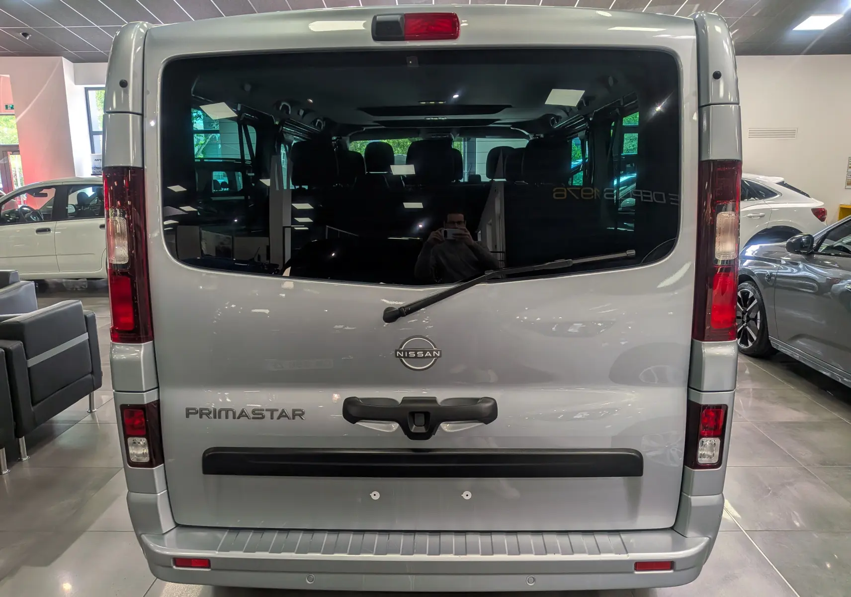 Vue arrière d'un Nissan Primastar Combi gris Highland 2026 avec vitre teintée et essuie-glace arrière visible en intérieur showroom.