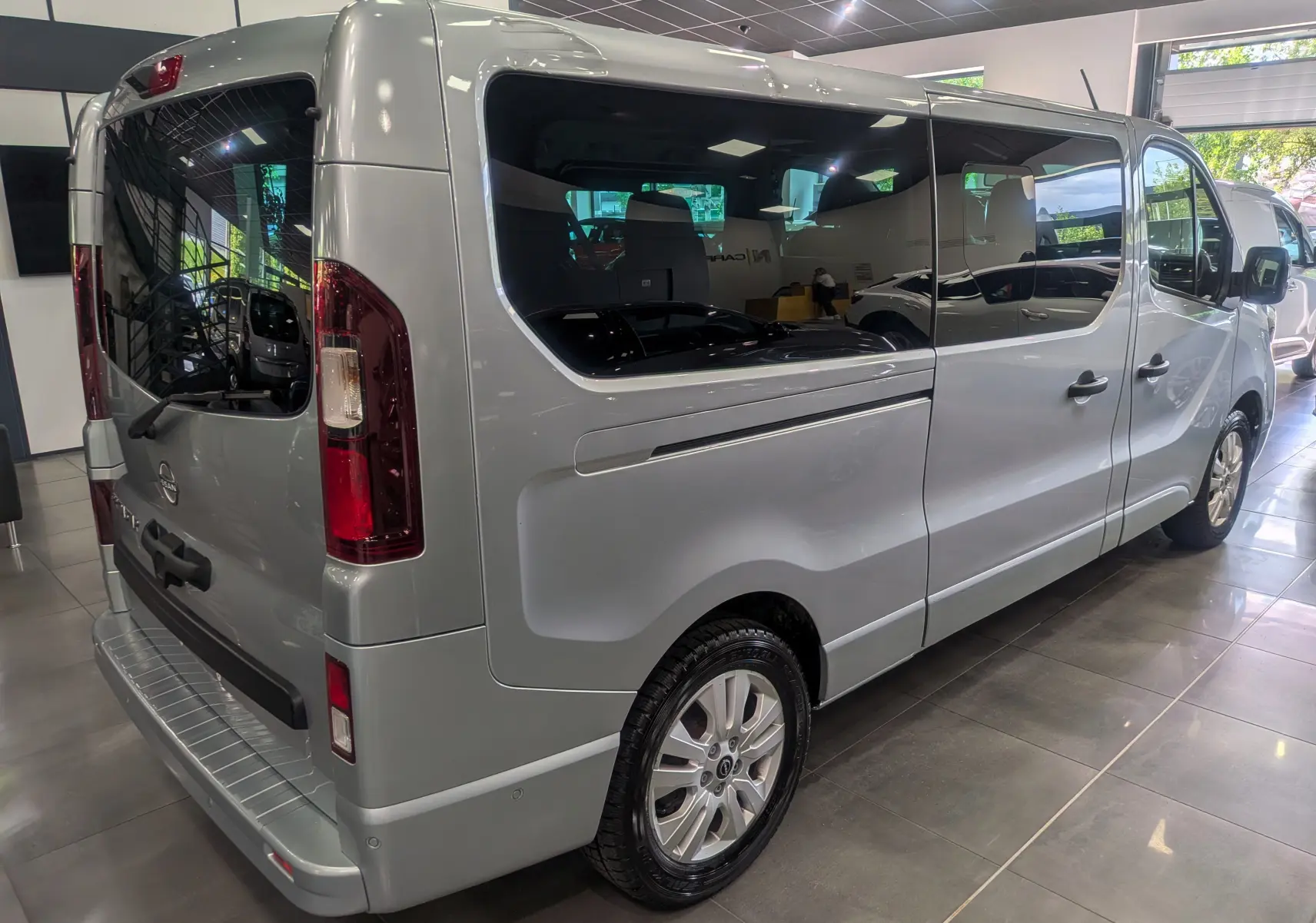 Vue 3/4 arrière droite du Nissan Primastar Combi gris Highland avec vitres teintées et jantes alliage en showroom.