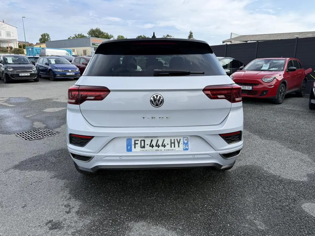 Vue arrière d'un Volkswagen T-Roc blanc avec toit noir et feux arrière LED, garé sur un parking extérieur.