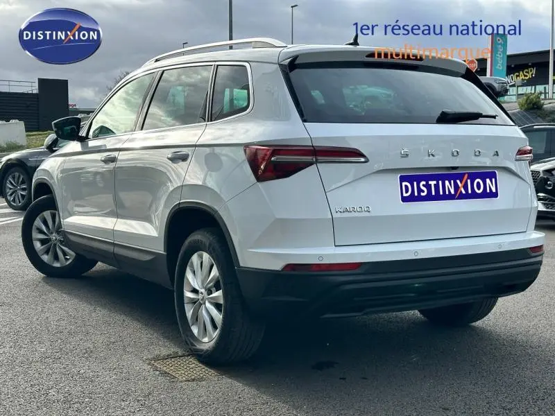 Vue 3/4 arrière droite d'un Skoda Karoq blanc lune métallisé avec barres de toit et jantes alu.