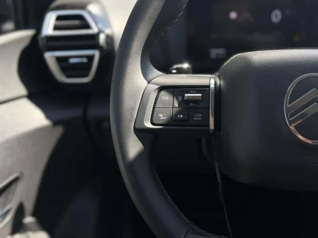 Gros plan sur la partie gauche du volant noir de la Citroën C4 Hybrid 145 ch 2025 avec commandes intégrées et logo centré.