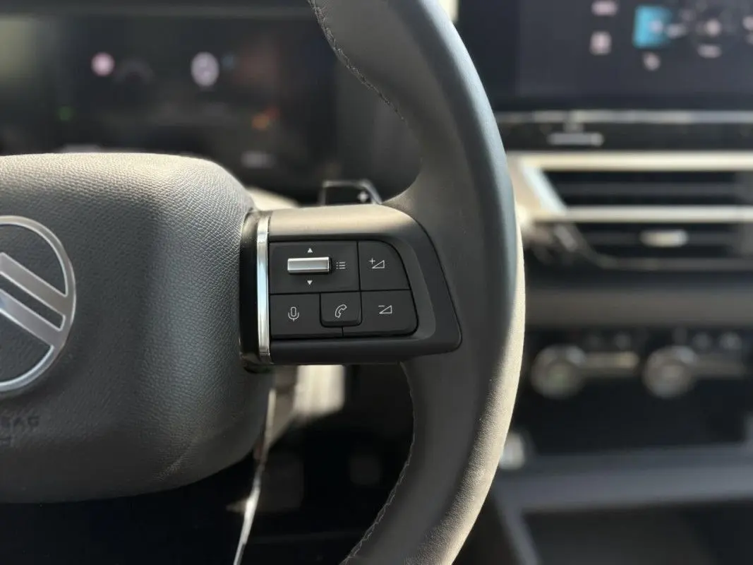 Gros plan sur les commandes gauche du volant noir de la Citroën C4 Hybrid 145 CH, avec tableau de bord flou en arrière-plan.