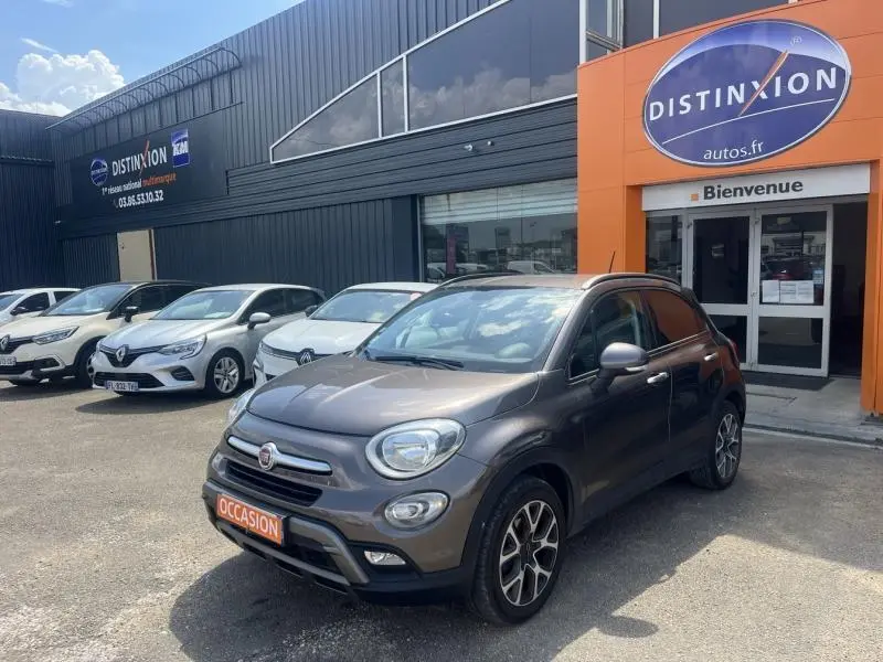 Fiat 500X bronze métallisé vue 3/4 avant droit, avec jantes alliage et calandre chromée, stationnée devant un concessionnaire.