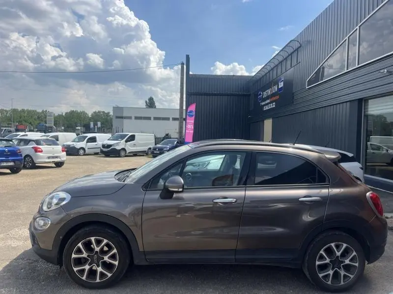Profil côté gauche d'un FIAT 500X bronze métallisé garé devant un bâtiment industriel sous un ciel partiellement nuageux.