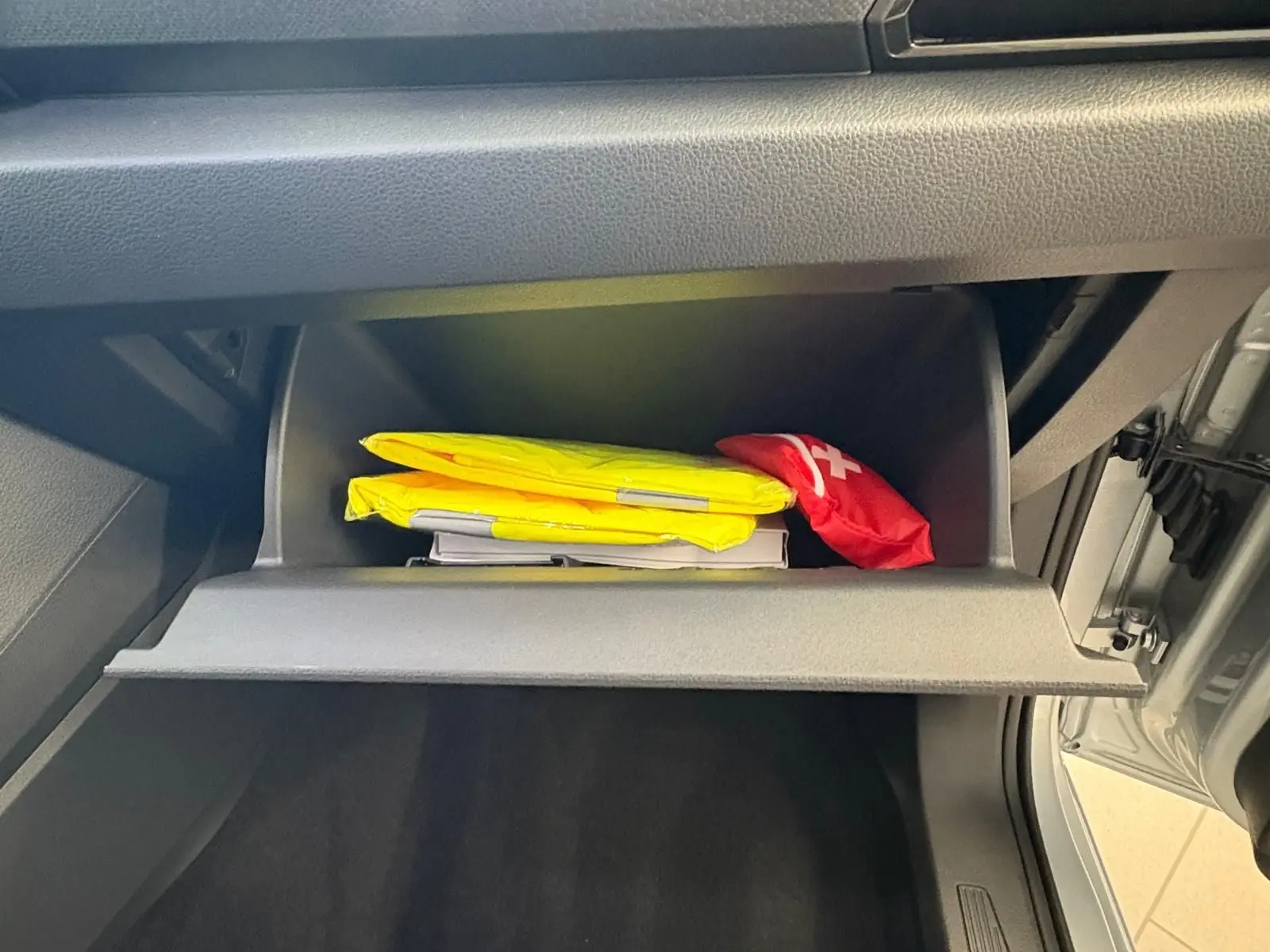 Compartiment de rangement ouvert côté passager du Volkswagen Caddy gris argent 2025, avec gilets de sécurité jaunes et trousse de secours rouge.