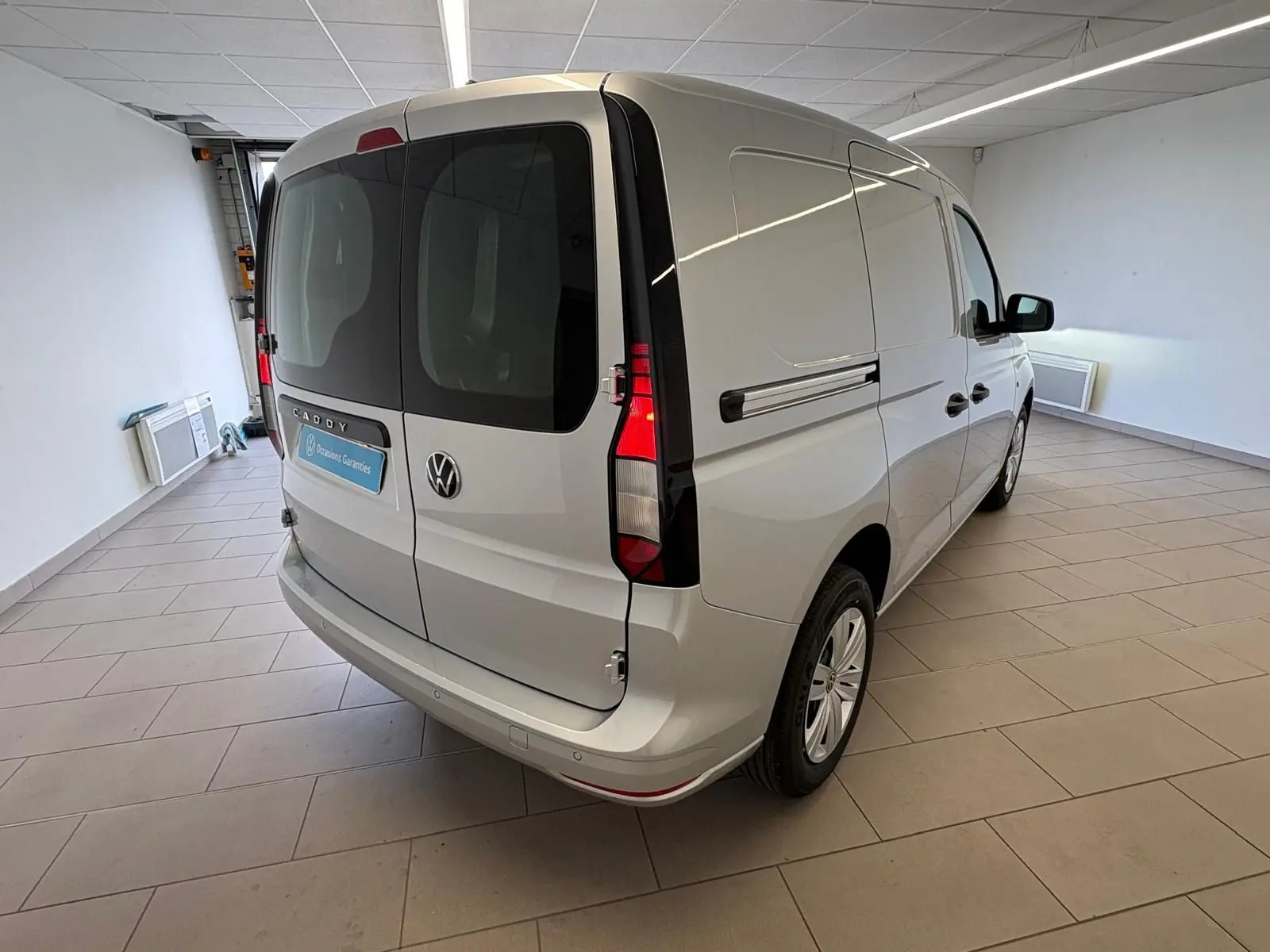 Vue 3/4 arrière droite d'un Volkswagen Caddy gris argent 2025 en version Business, avec portes arrière vitrées et feux verticaux.