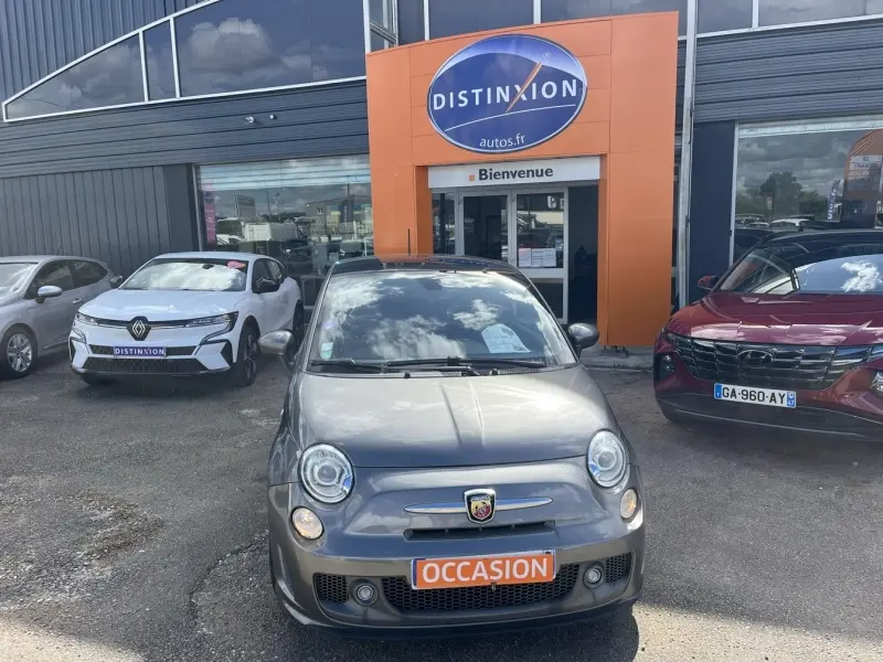 Vue avant d'une ABARTH 500 gris foncé métal avec badge Abarth et plaque orange "OCCASION" devant un garage.
