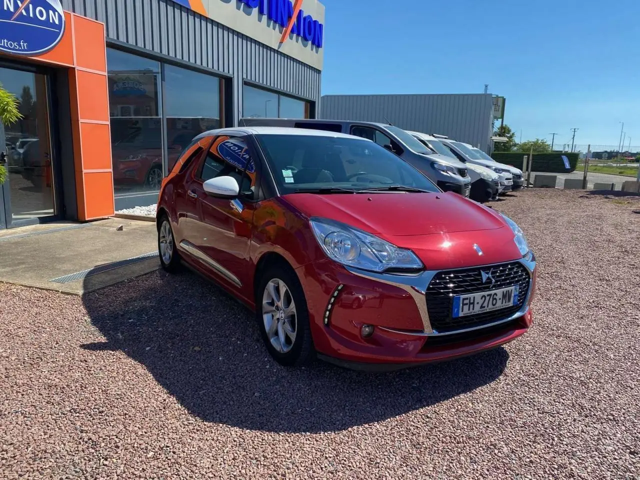 DS3 rouge avec toit blanc, vue 3/4 avant droit, garée devant un bâtiment commercial sous un ciel bleu.