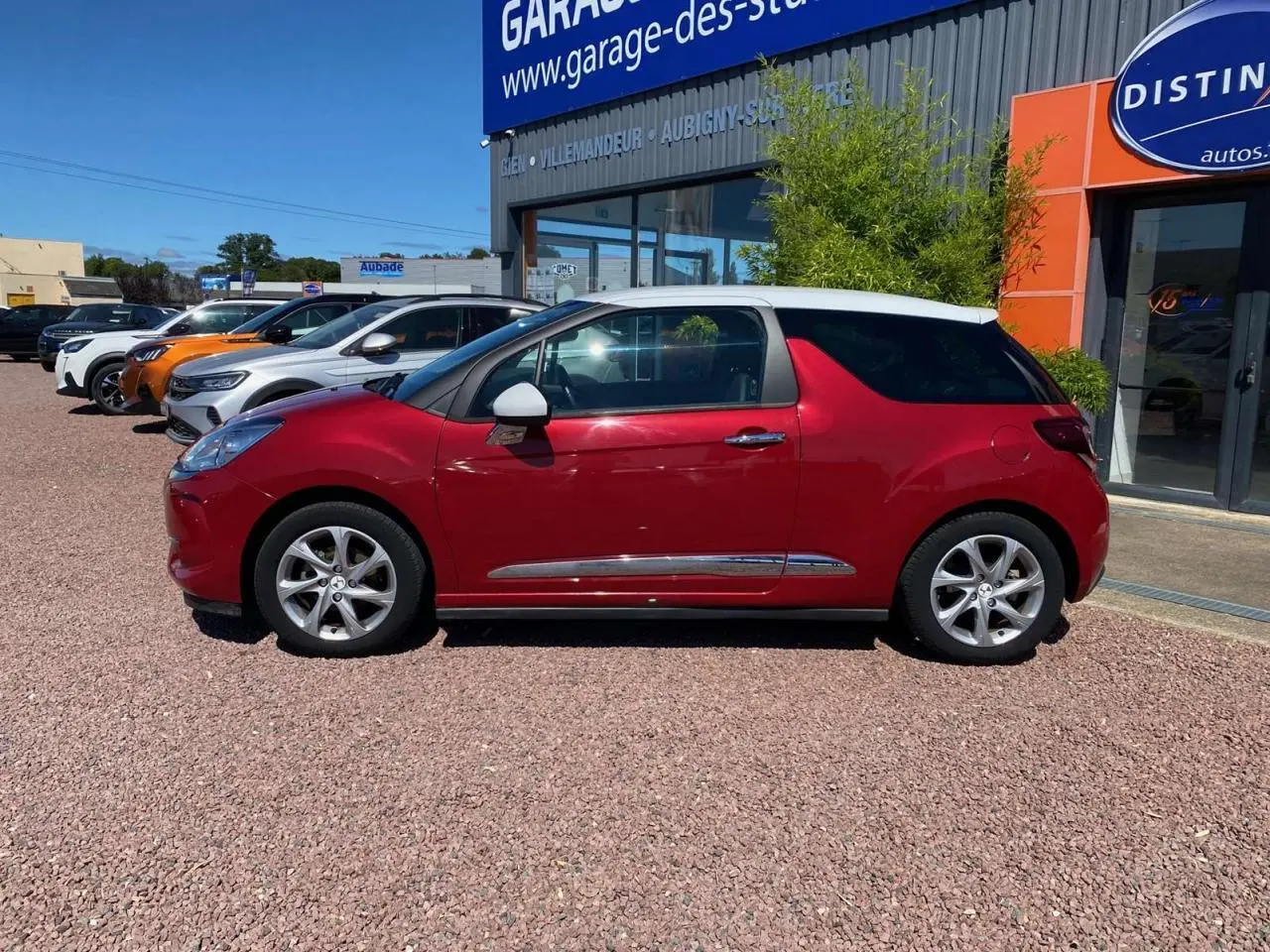 Profil côté gauche d'une DS3 rouge avec toit blanc, jantes alliage 16 pouces, stationnée devant un garage.