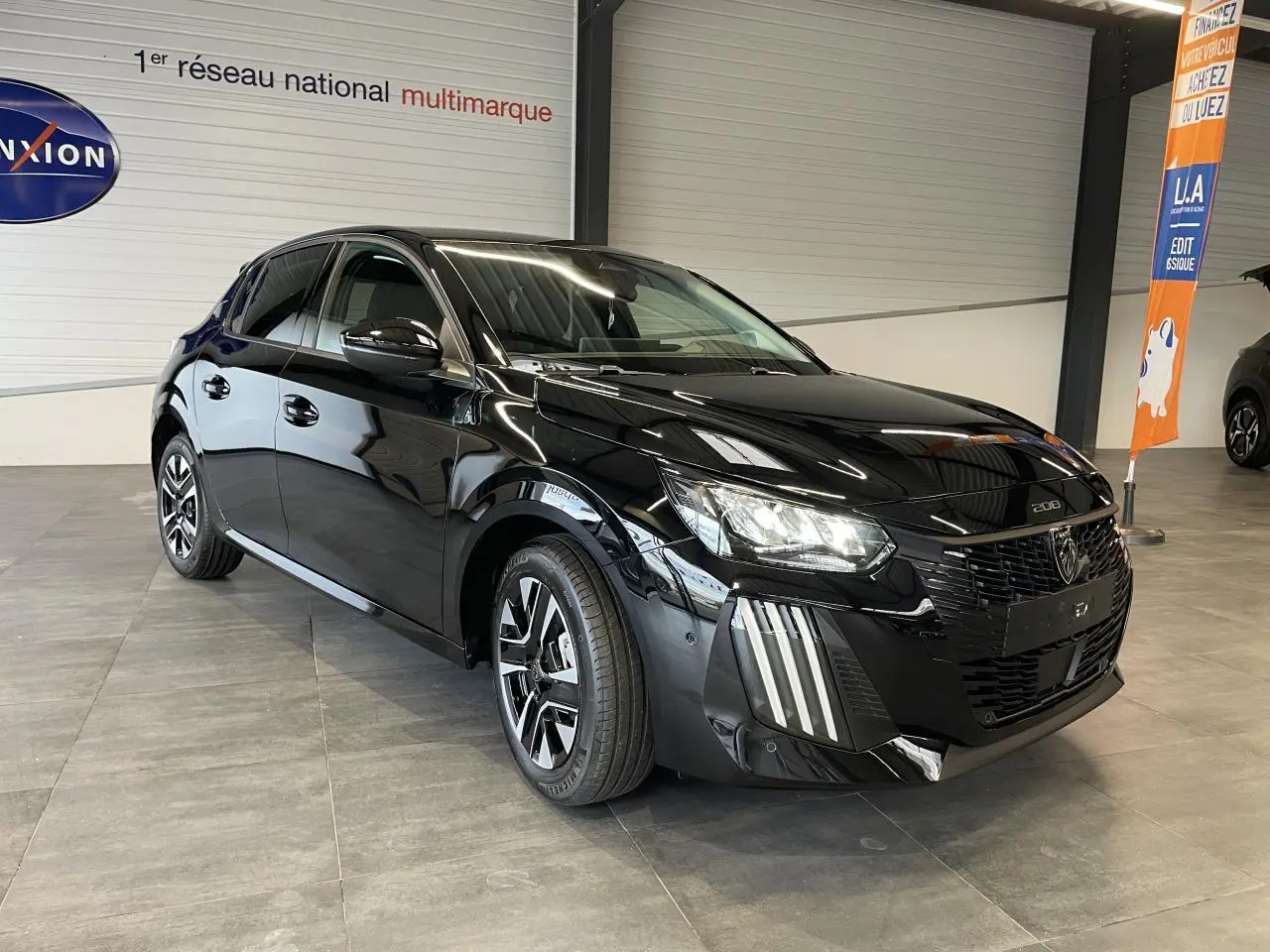 Peugeot 208 noire vue 3/4 avant droit en intérieur, avec jantes alliage bi-tons et feux LED distinctifs.