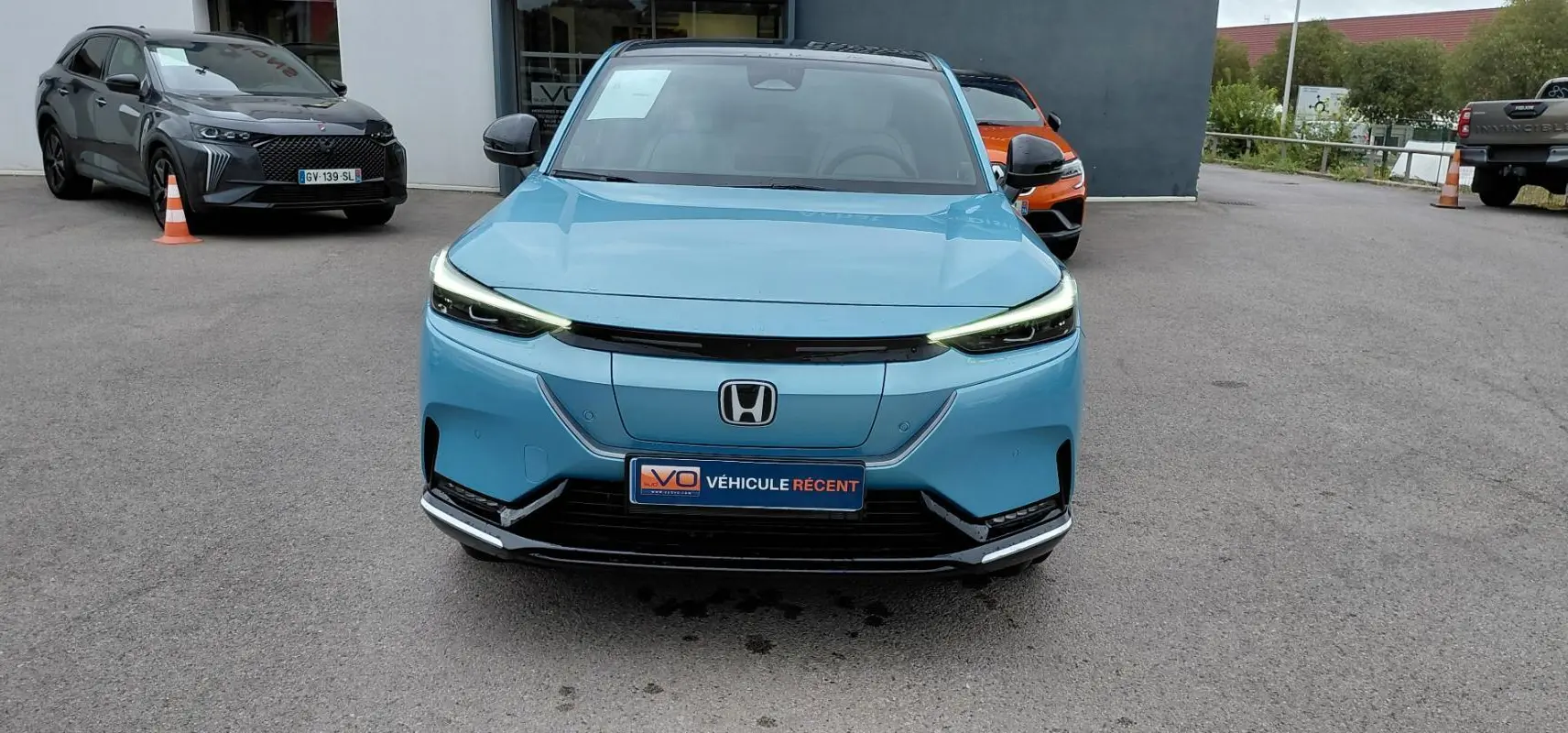 Vue frontale d'une Honda e:Ny1 Advance 204 ch 2025 en bleu électrique avec phares LED allumés sur parking.