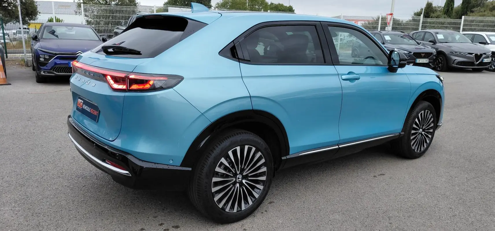 Vue 3/4 arrière droite d'un Honda e:Ny1 Advance bleu électrique 2025 avec feux arrière allumés et jantes stylisées.