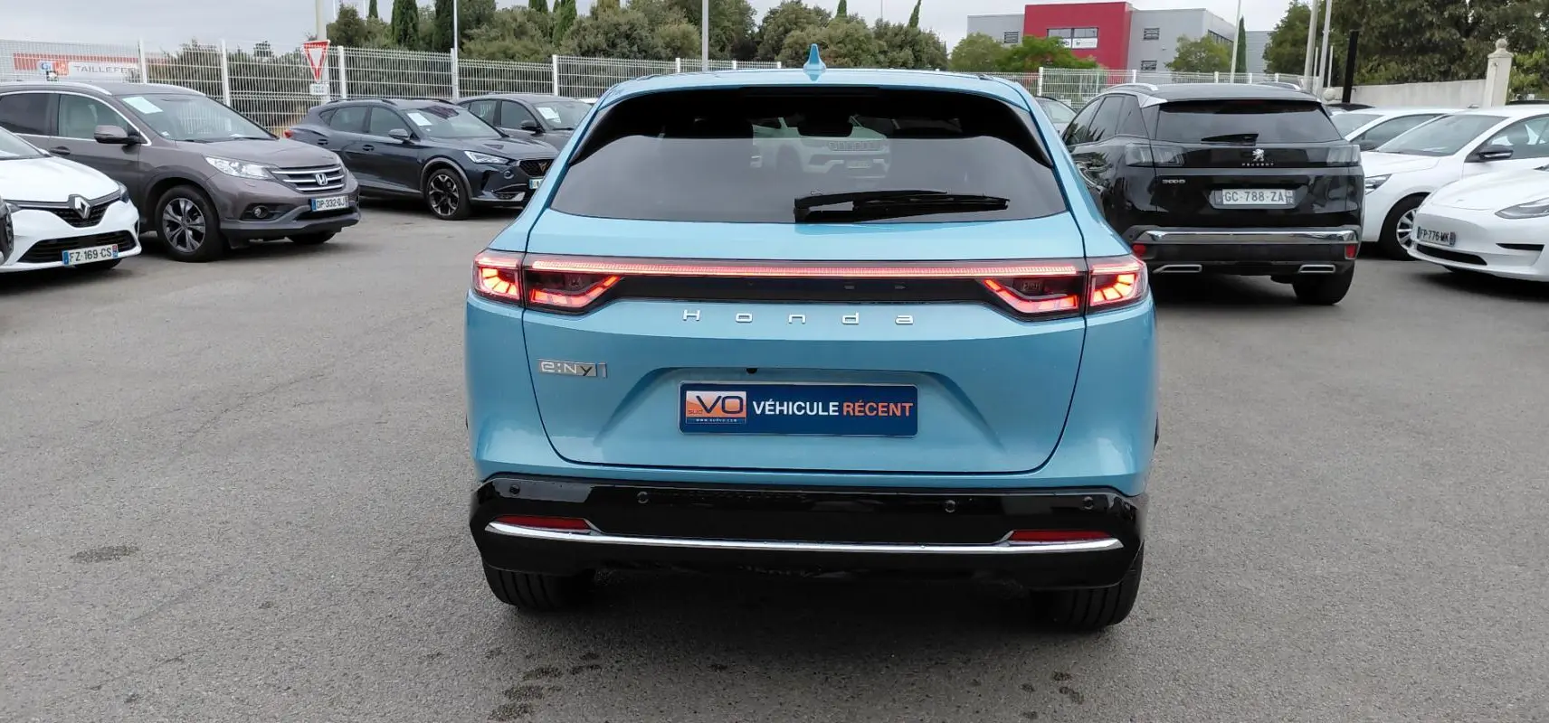 Vue arrière d'un Honda e:Ny1 bleu 2025 avec feux LED allumés et logo Honda visible sur un parking.