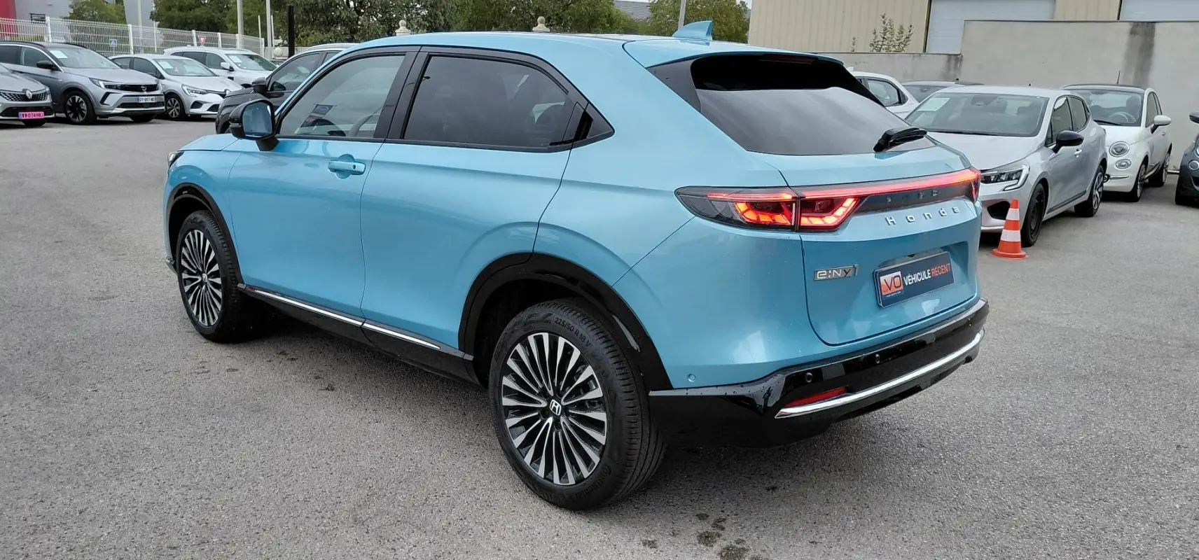 Vue 3/4 arrière droite d’un Honda e:Ny1 bleu électrique 2025 avec feux arrière allumés et jantes stylisées.
