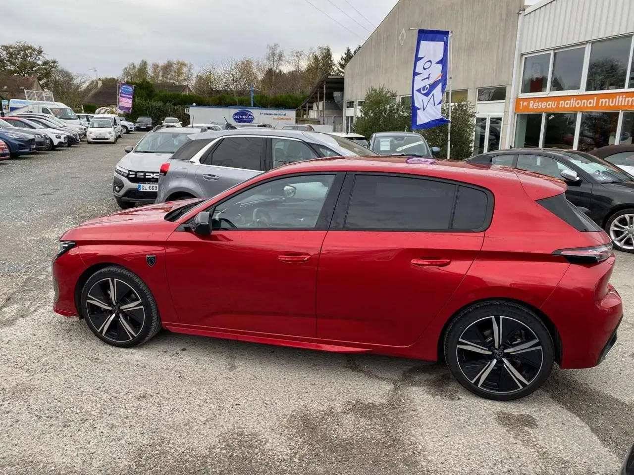 Profil latéral droit de la Peugeot 308 GT 2022 rouge Elixir avec jantes noires et vitres teintées sur parking extérieur.