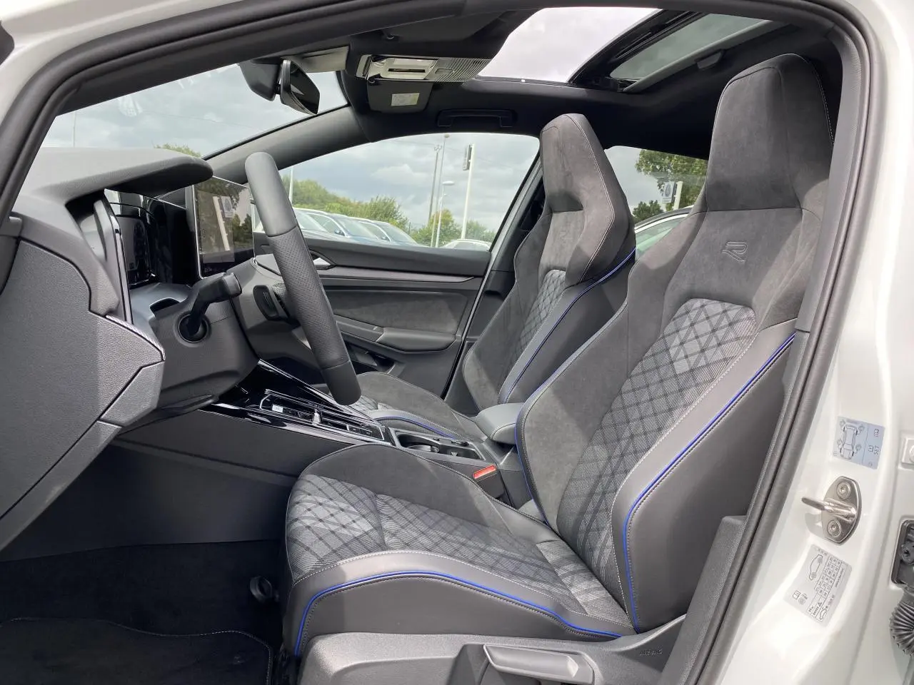 Intérieur côté conducteur d'une Volkswagen Golf 1.5 eTSI R-Line blanche, sièges sport gris avec surpiqûres bleues et toit panoramique.
