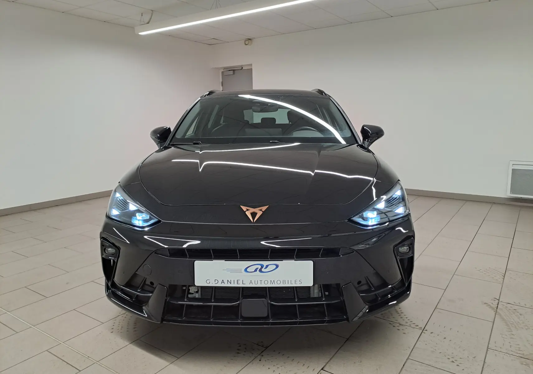 Vue frontale d'une CUPRA LEON ST 1.5 eTSI 150 DSG7 en Noir Minuit avec phares allumés et logo cuivré distinctif.