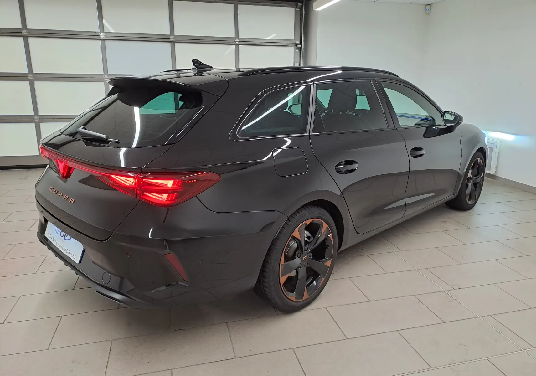 Vue 3/4 arrière droite de la CUPRA LEON ST 1.5 eTSI 150 DSG7 en Noir Minuit avec jantes cuivrées et feux arrière allumés.