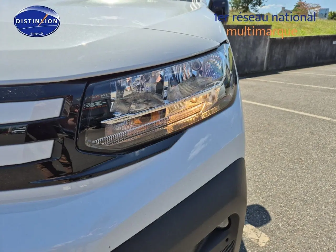 Gros plan sur le phare avant droit du Peugeot Partner 2025 blanc, avec calandre noire et détails lumineux allumés.