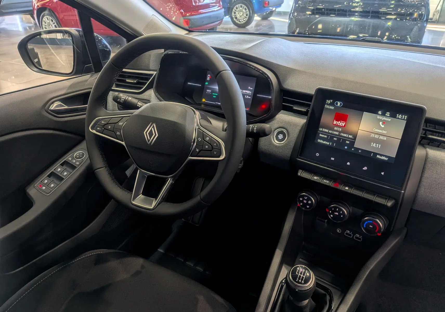 Vue intérieure côté conducteur de la Renault Clio V 2025, tableau de bord moderne avec écran tactile et volant multifonction noir.