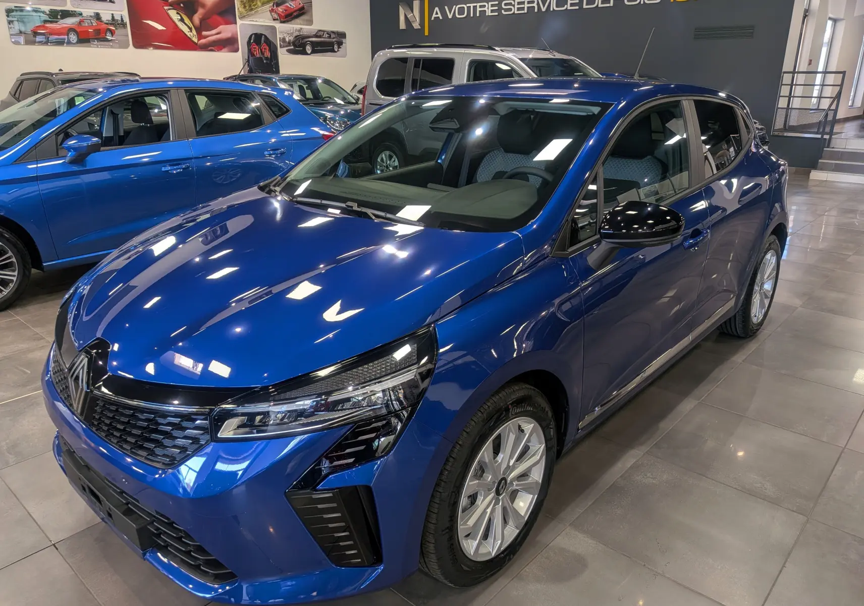 Renault Clio V Bleu Iron vue 3/4 avant droit en showroom, avec jantes alliage et rétroviseurs noirs.