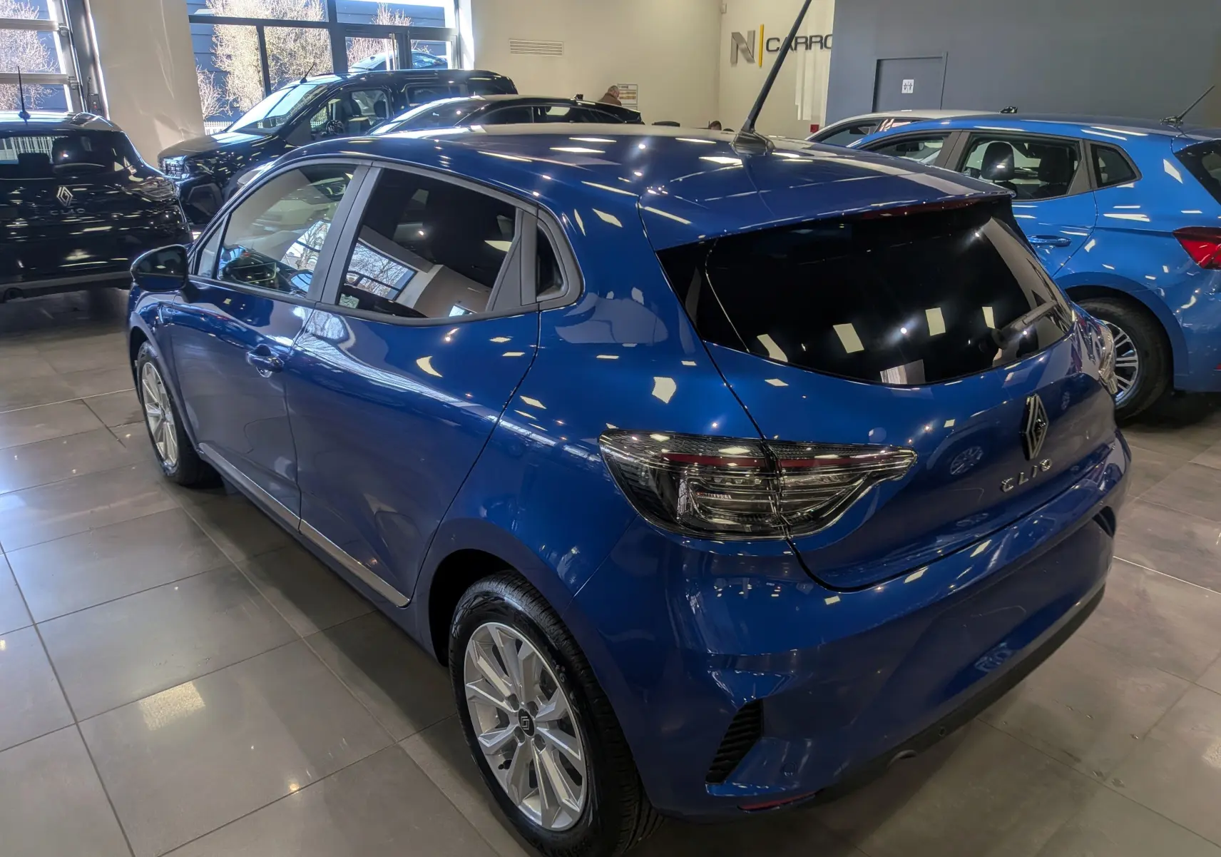Vue 3/4 arrière droite d'une Renault Clio V Bleu Iron 2025 en showroom, avec vitres teintées et jantes alliage.