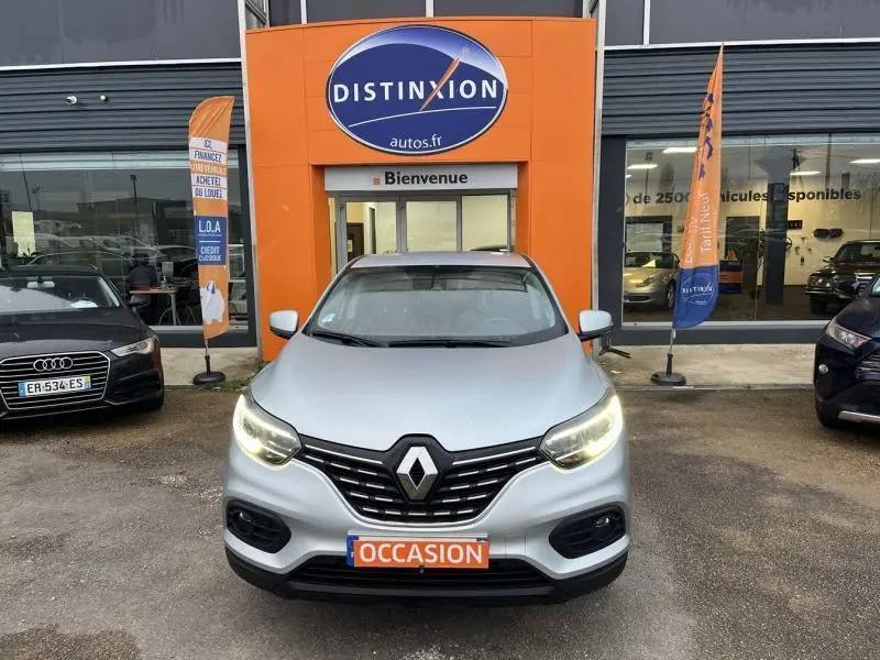 Vue avant d'un Renault Kadjar blanc glacier avec phares allumés et plaque "OCCASION" devant un concessionnaire Distinxion.