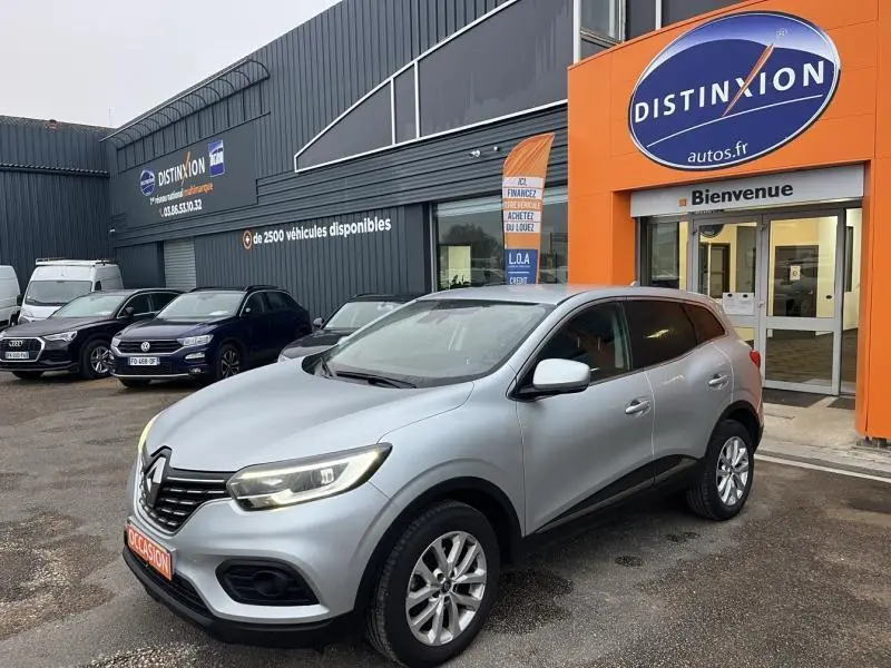 Renault Kadjar blanc en 3/4 avant droit stationné devant un concessionnaire avec panneaux orange et gris.