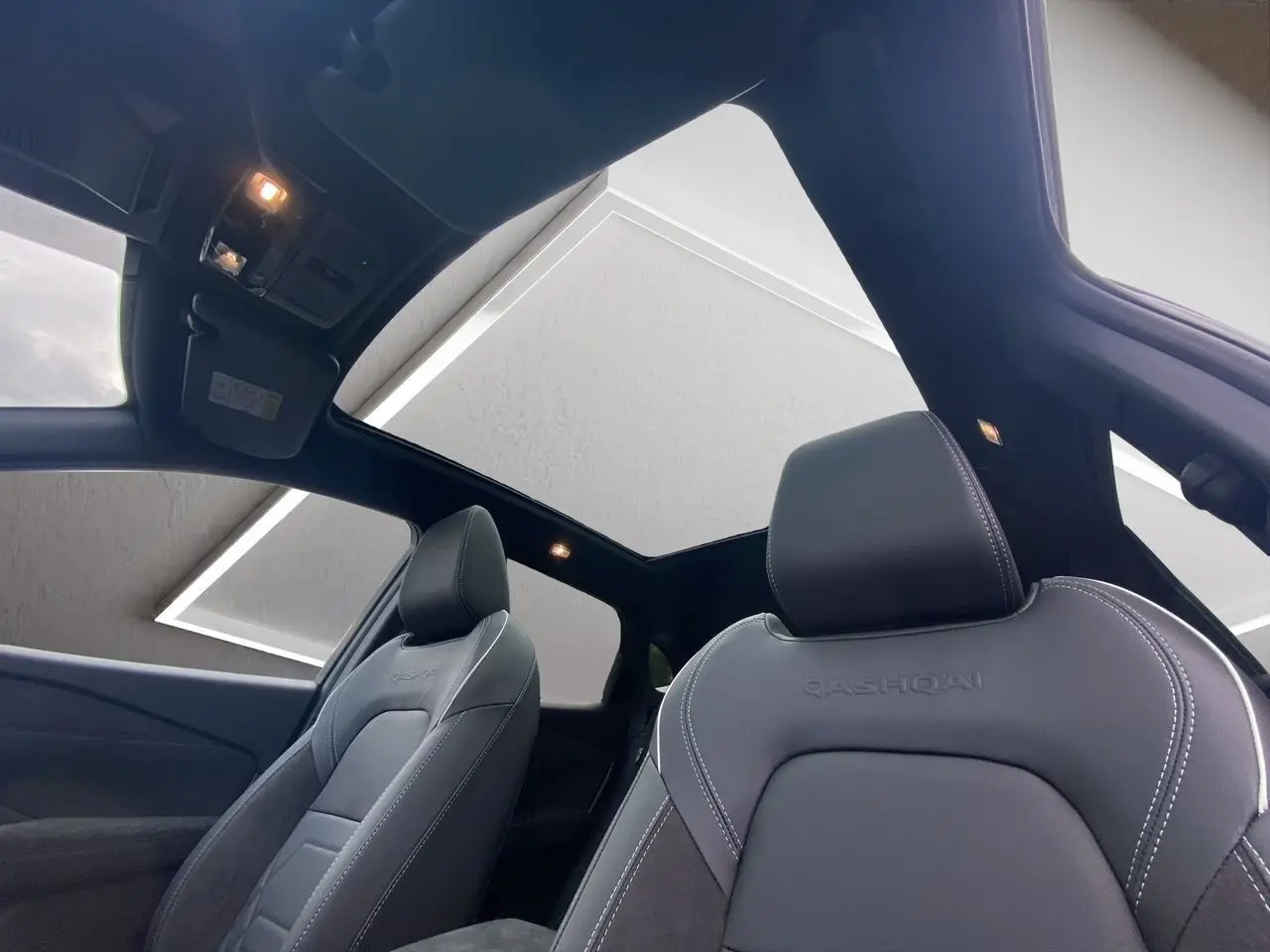 Vue intérieure du Nissan Qashqai 2025 montrant les sièges avant noirs avec surpiqûres blanches et toit panoramique.