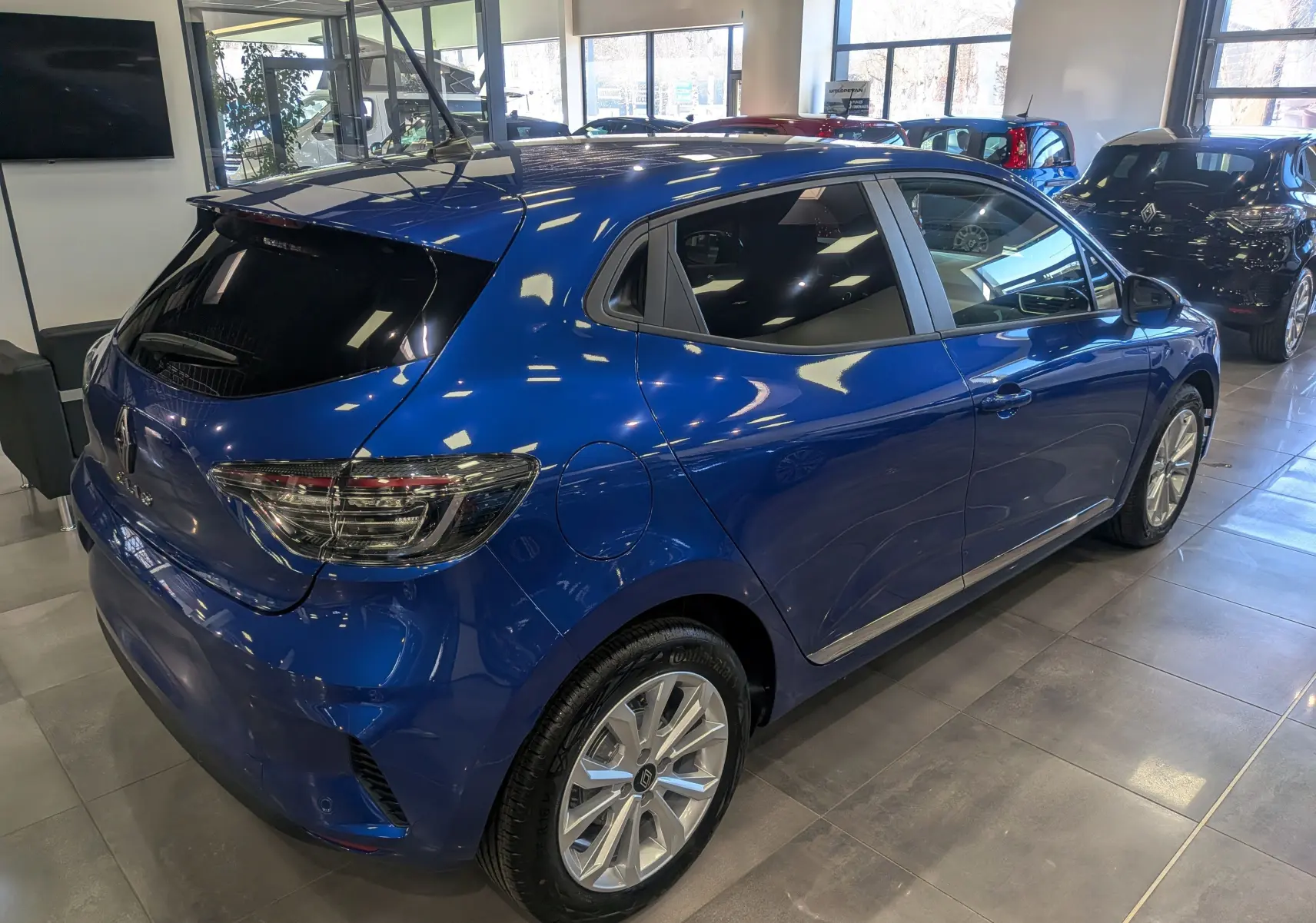 Vue 3/4 arrière droite d'une Renault Clio V Bleu Iron 2025 en intérieur, avec jantes alliage et vitres teintées.