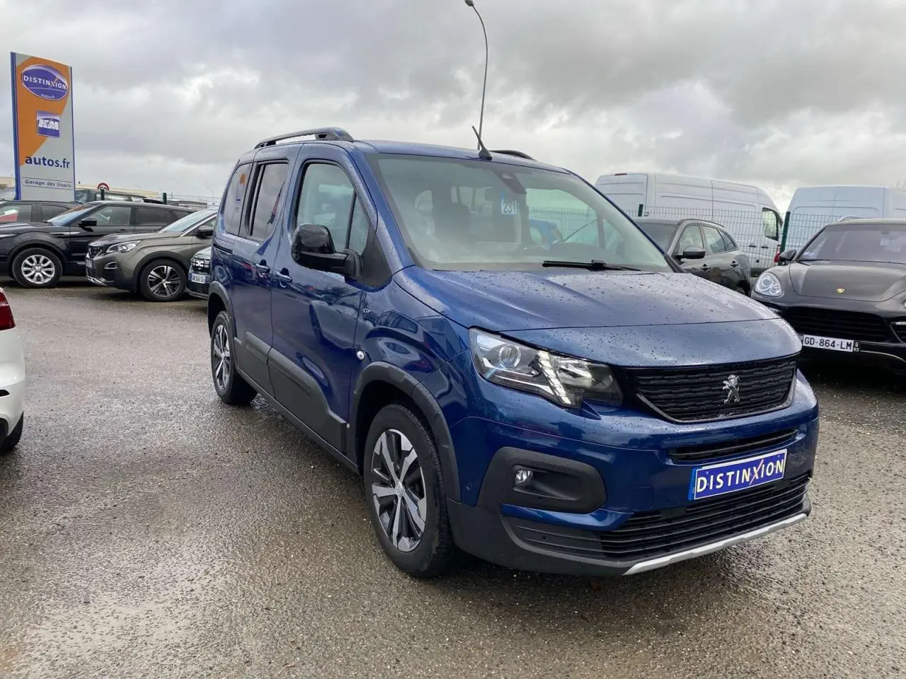 Peugeot Rifter GT bleu métallisé en 3/4 avant droit sous ciel nuageux sur parking humide.