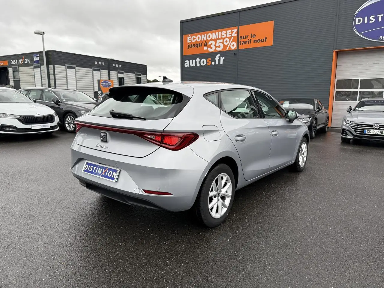 Vue 3/4 arrière droite d'une SEAT Leon 2022 en argent urban, stationnée devant un concessionnaire sous un ciel nuageux.